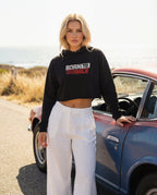 Woman wearing a black 'BORN TO RACE' hoodie and white pants standing next to a vintage car.